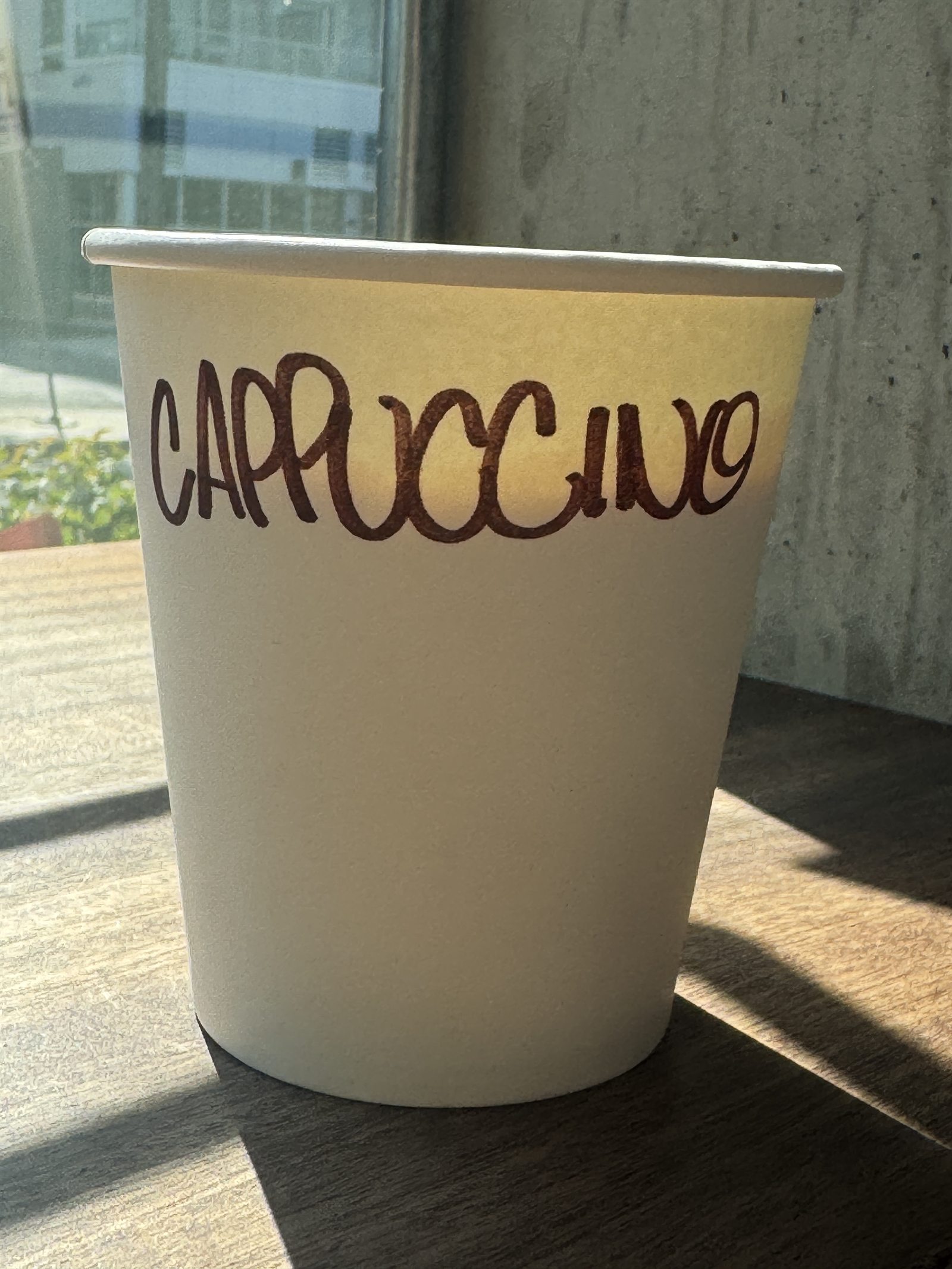 A paper cup on a sunlit table, CAPPUCCINO written across it in brown marker.